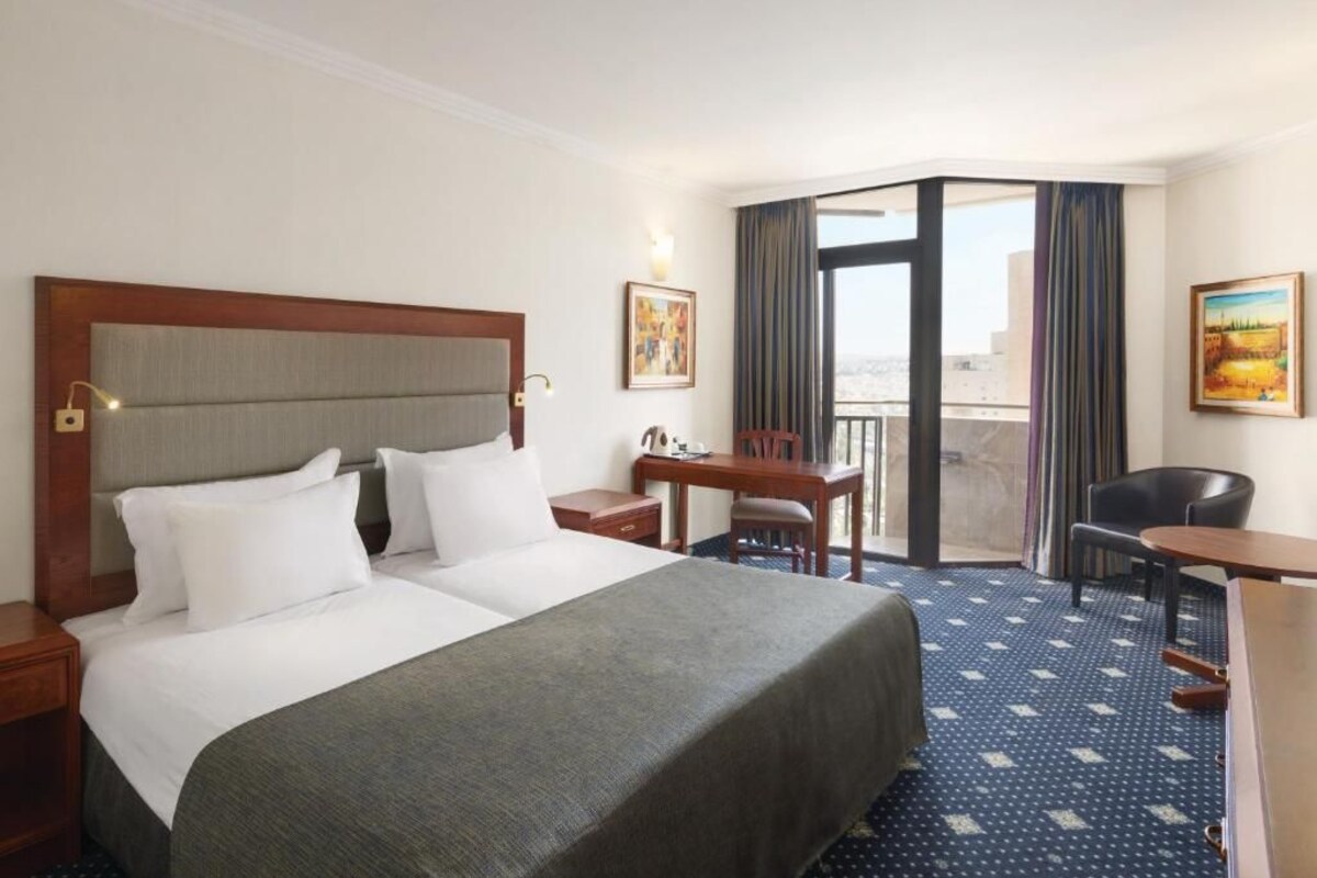 A hotel room with a large bed, a desk, two chairs, a small table, a balcony with curtains, and a painting on the wall.