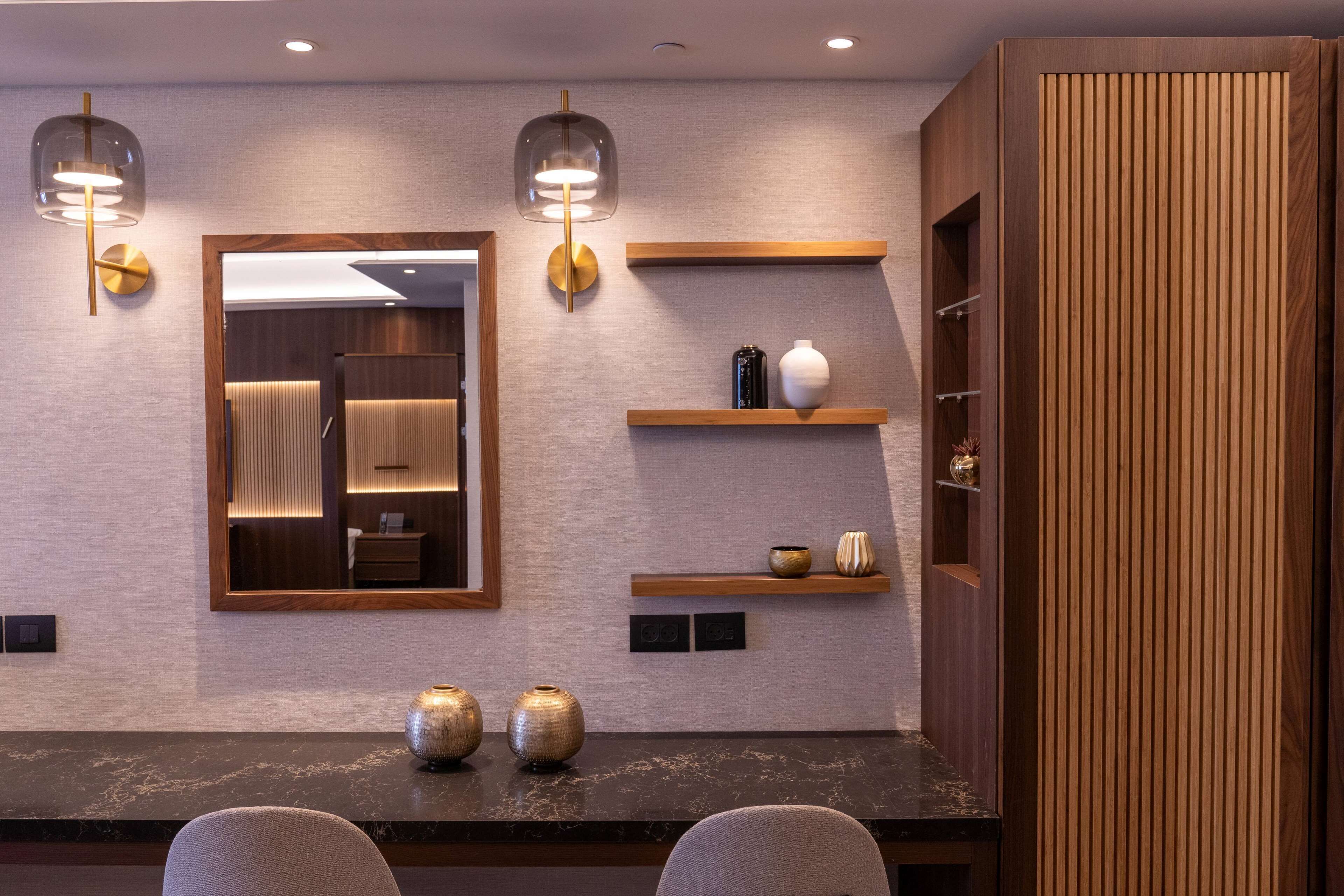 A modern hotel lobby with a marble reception desk, two seating areas, a large mirror, wall-mounted shelves with decorative items, and a wooden panel wall.
