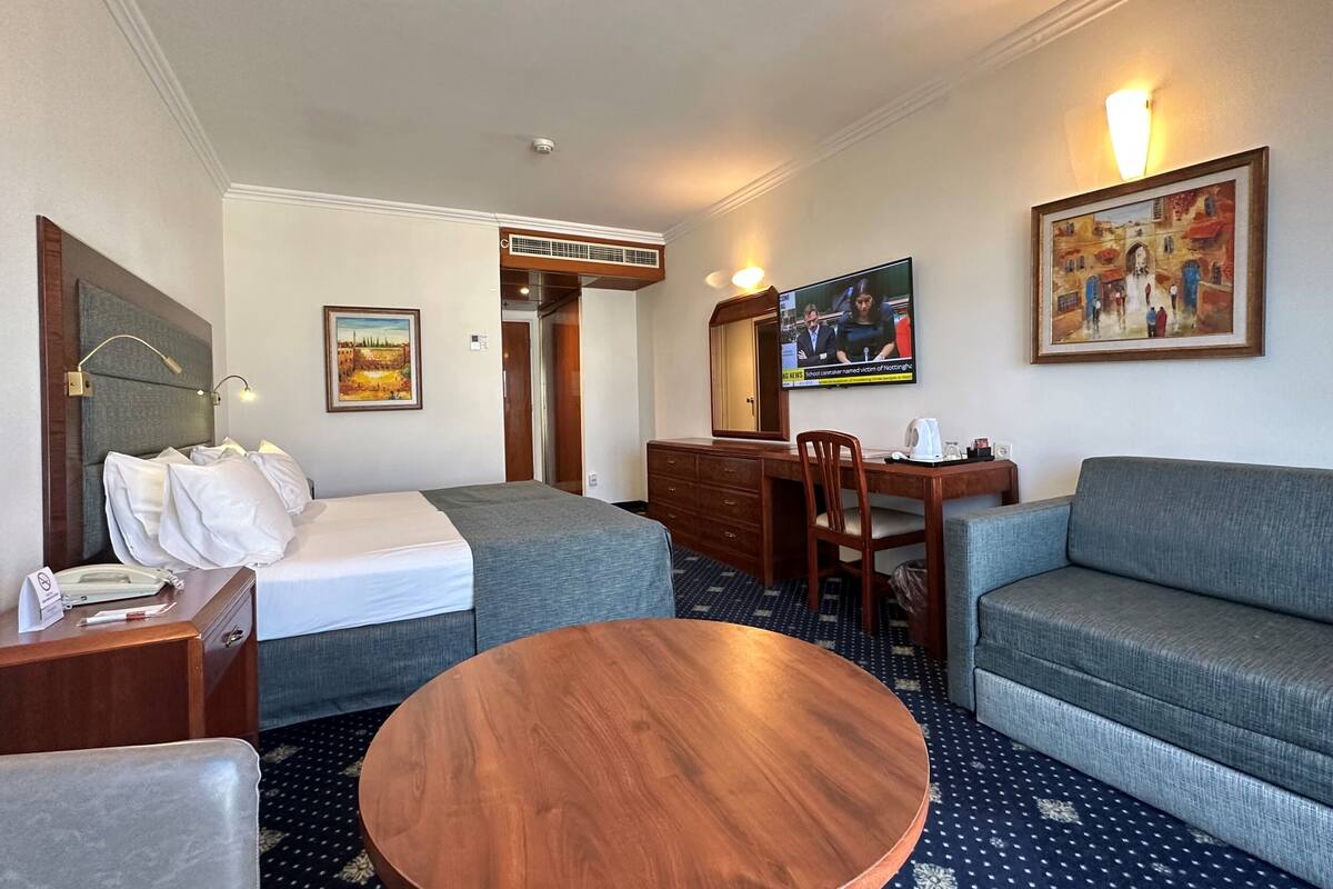 Quarto de hotel com cama, sofá, mesa de madeira redonda, escrivaninha com cadeira, televisão e um quadro na parede.