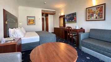 Een hotelkamer met een bed, een slaapbank, een ronde houten tafel, een bureau met stoel, een televisie en een schilderij aan de muur.