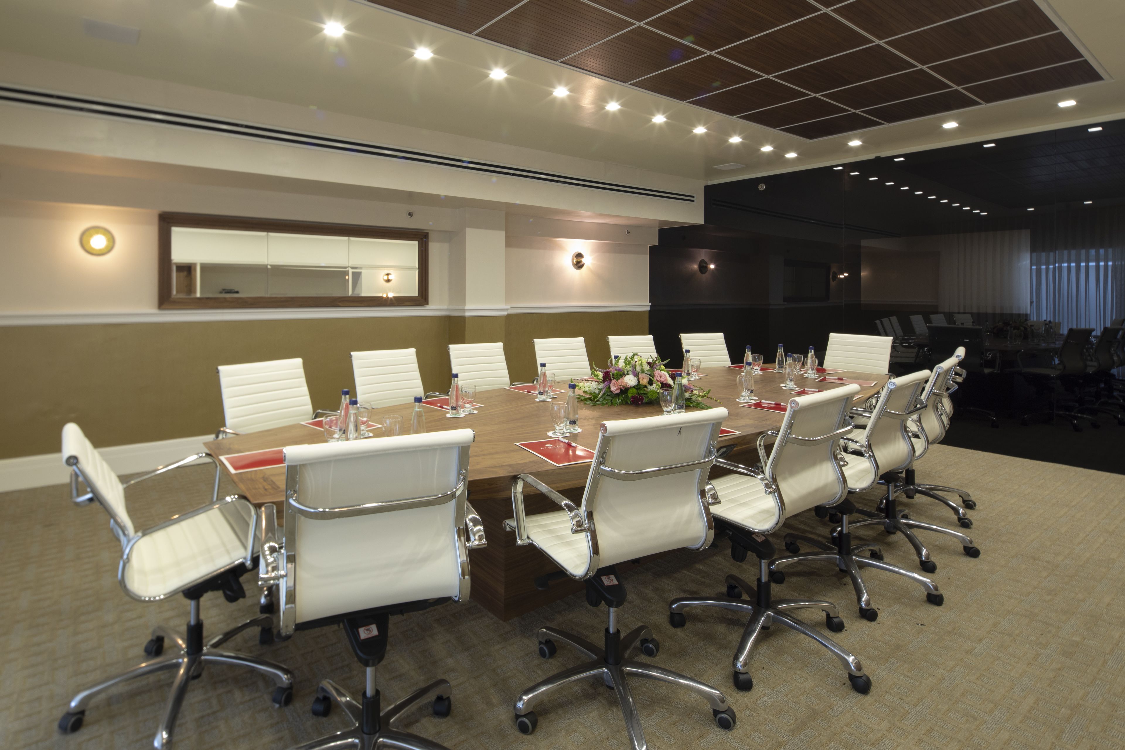 A conference room with a long table, white chairs, and a large mirror on the wall.