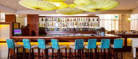 Un espace bar avec un comptoir en bois, de nombreuses bouteilles d’alcool et des tabourets de bar bleus à motifs.
