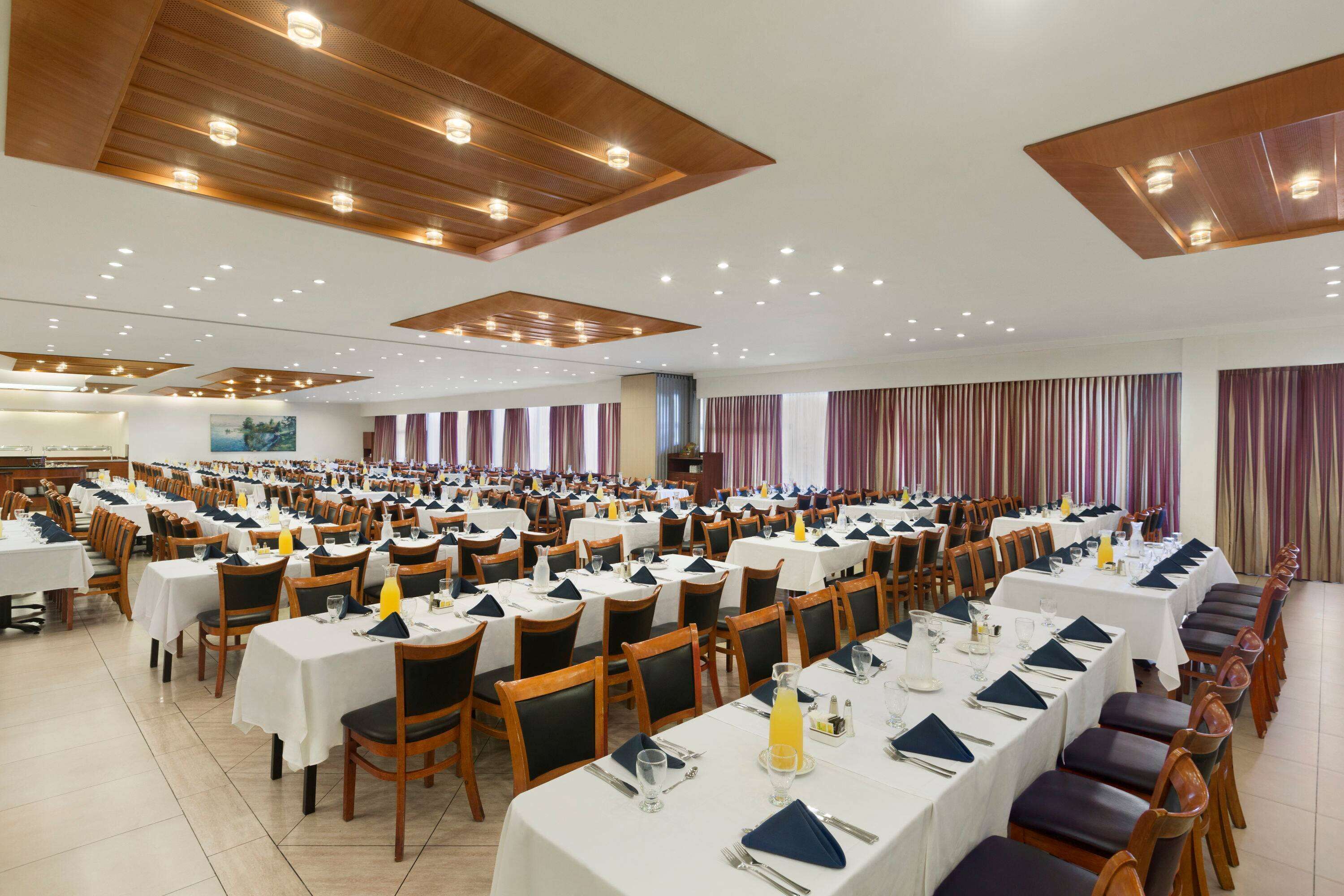 A large banquet hall with rows of tables set for dining, wooden ceiling panels, and recessed lighting.