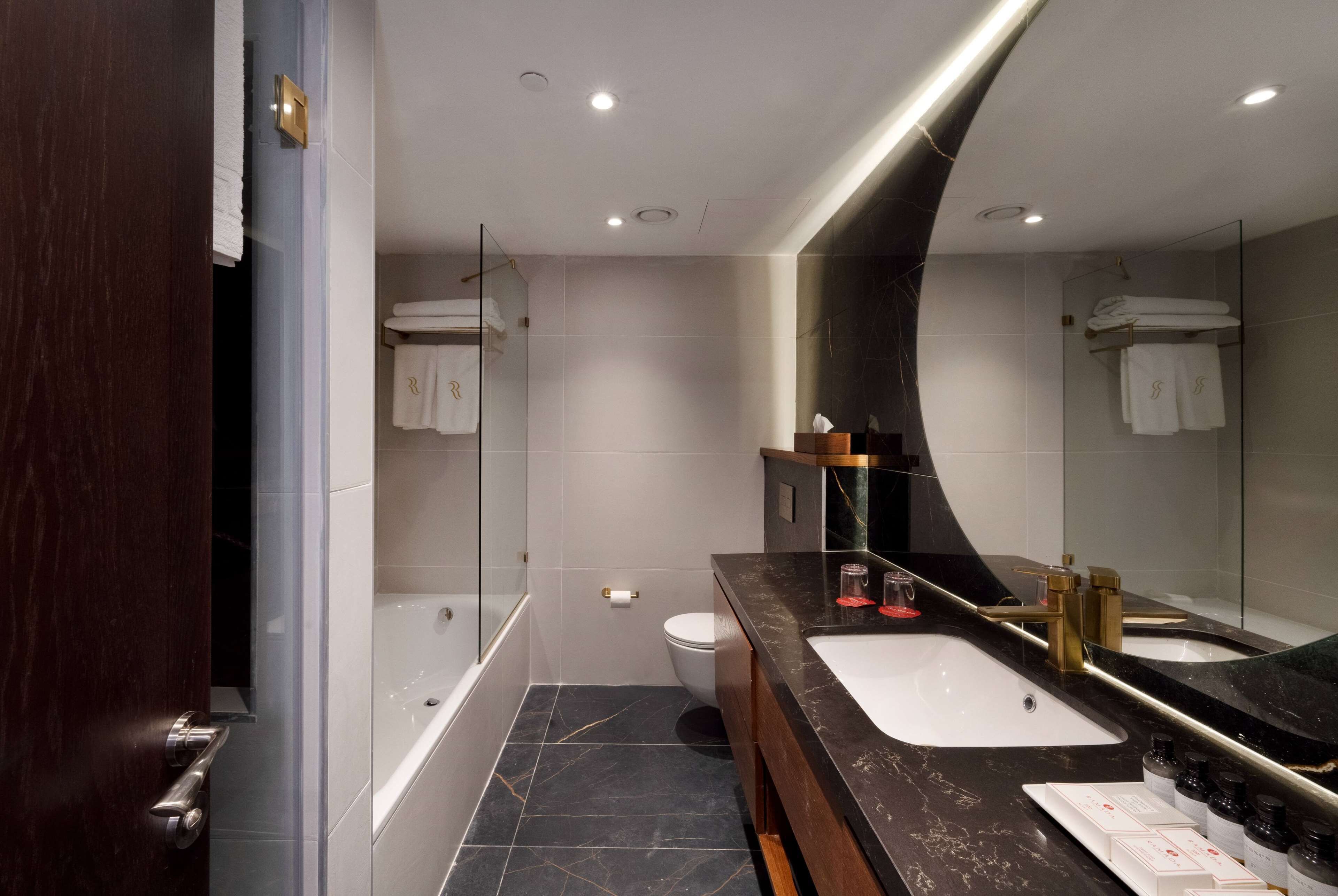 A modern bathroom with a large mirror, two sinks, a bathtub, and a toilet.