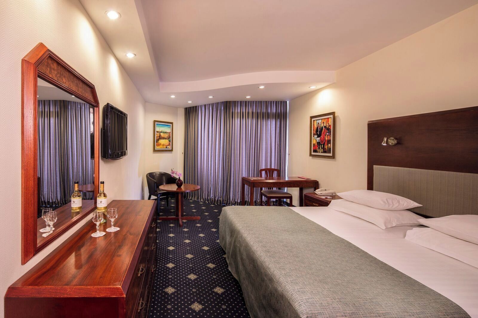 Une chambre d’hôtel comprenant un lit, un bureau avec une chaise, un miroir, une télévision et une fenêtre avec des rideaux.