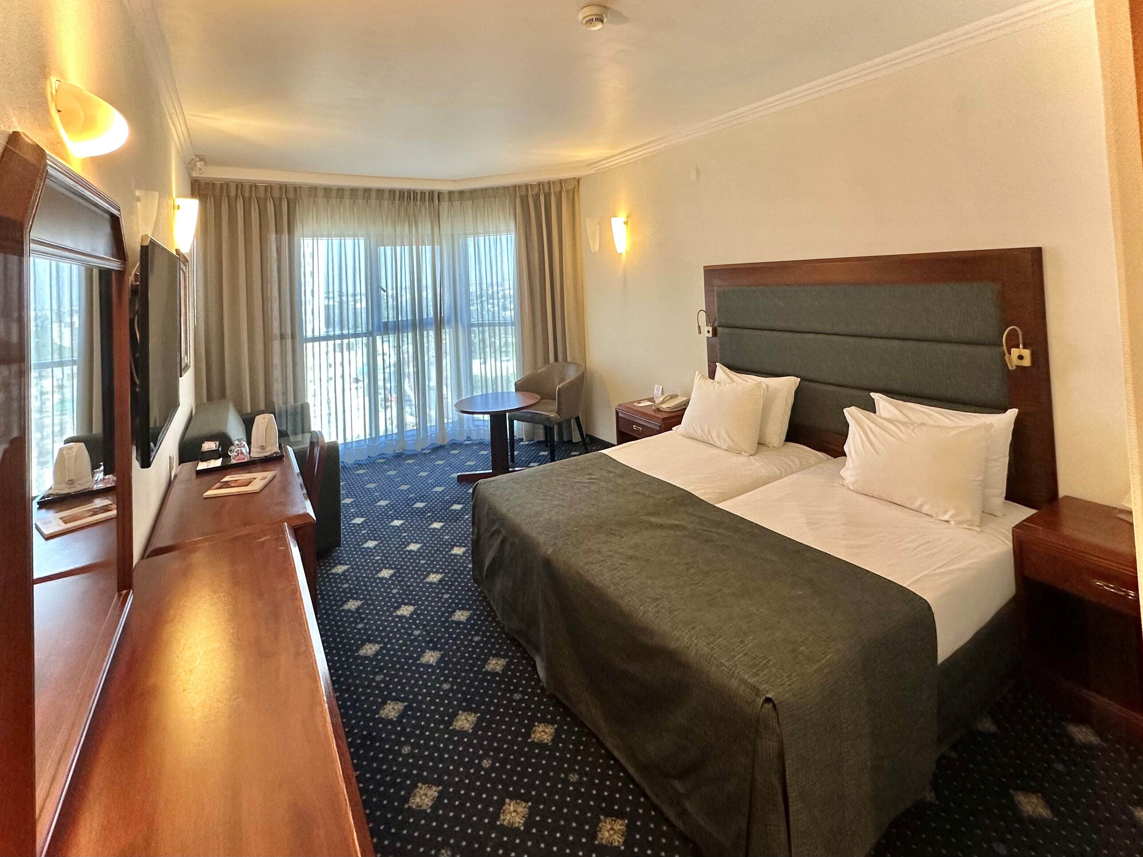 A hotel room with a large bed, a desk, a chair, a small table, and a window with curtains.
