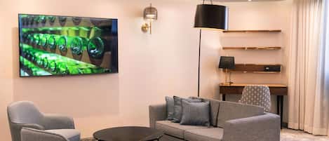 Un soggiorno moderno con un divano grigio, due poltrone, un tavolino, una TV a schermo piatto che mostra bottiglie di vetro verde, una lampada da terra e una scrivania con scaffali.