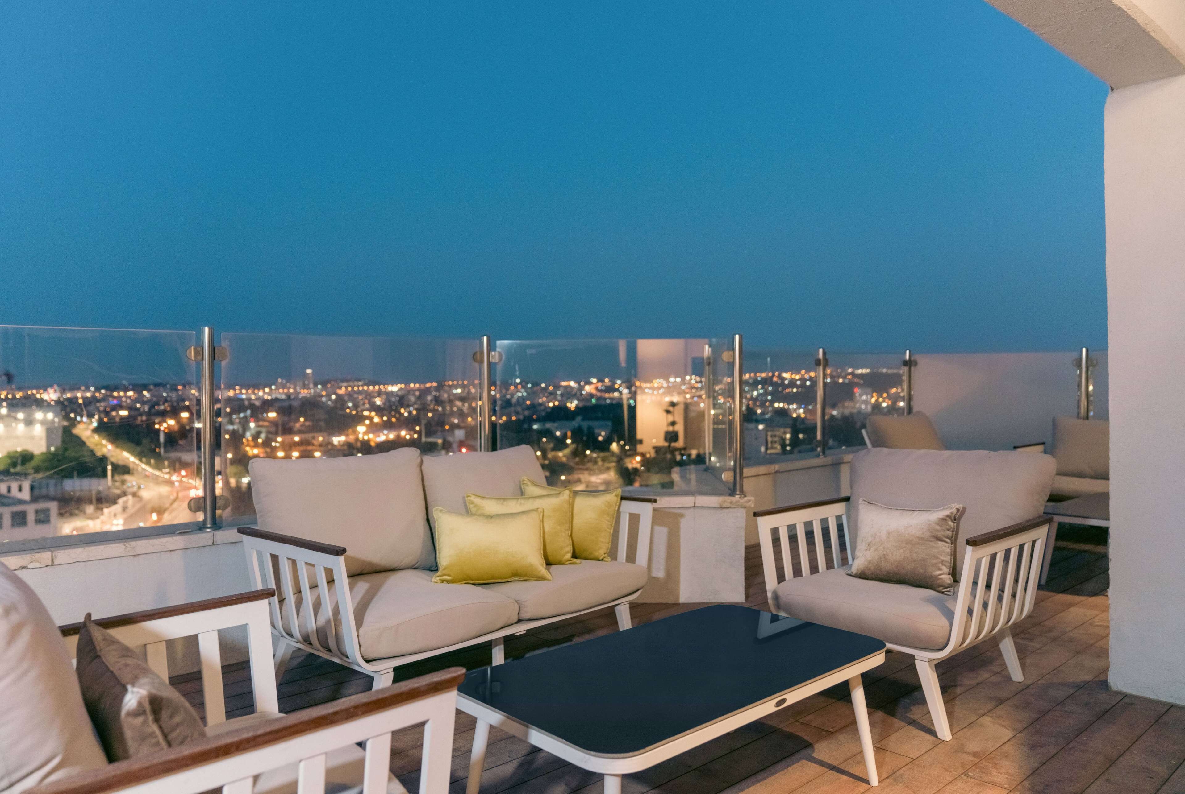 A balcony with a sofa and chairs, a small table, and a city view at dusk.