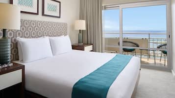 A hotel room with a large bed, two bedside lamps, a view of the ocean through a sliding door, and a balcony with chairs and tables.