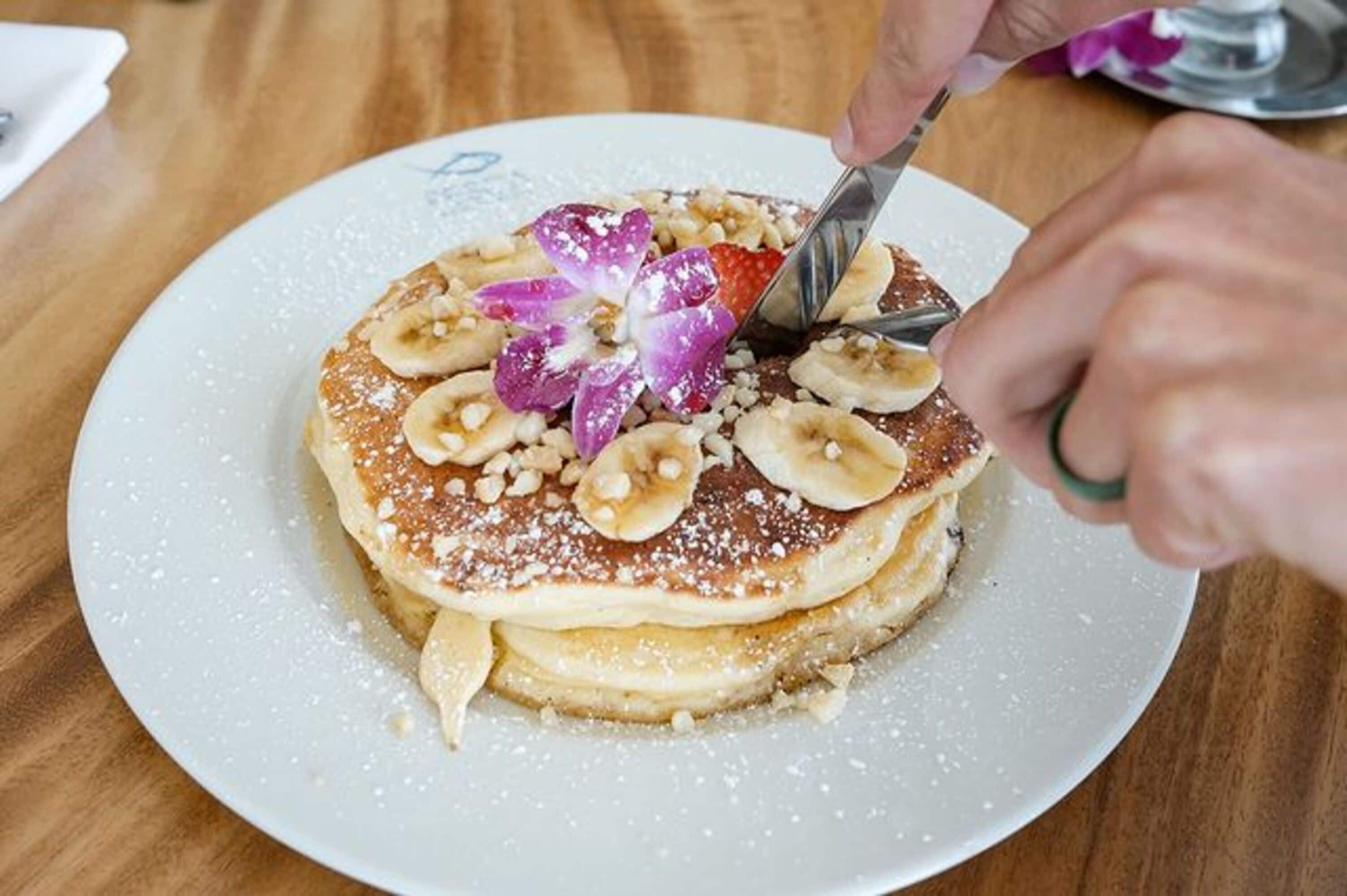 A stack of pancakes with bananas and a purple flower on top, being cut with a knife.