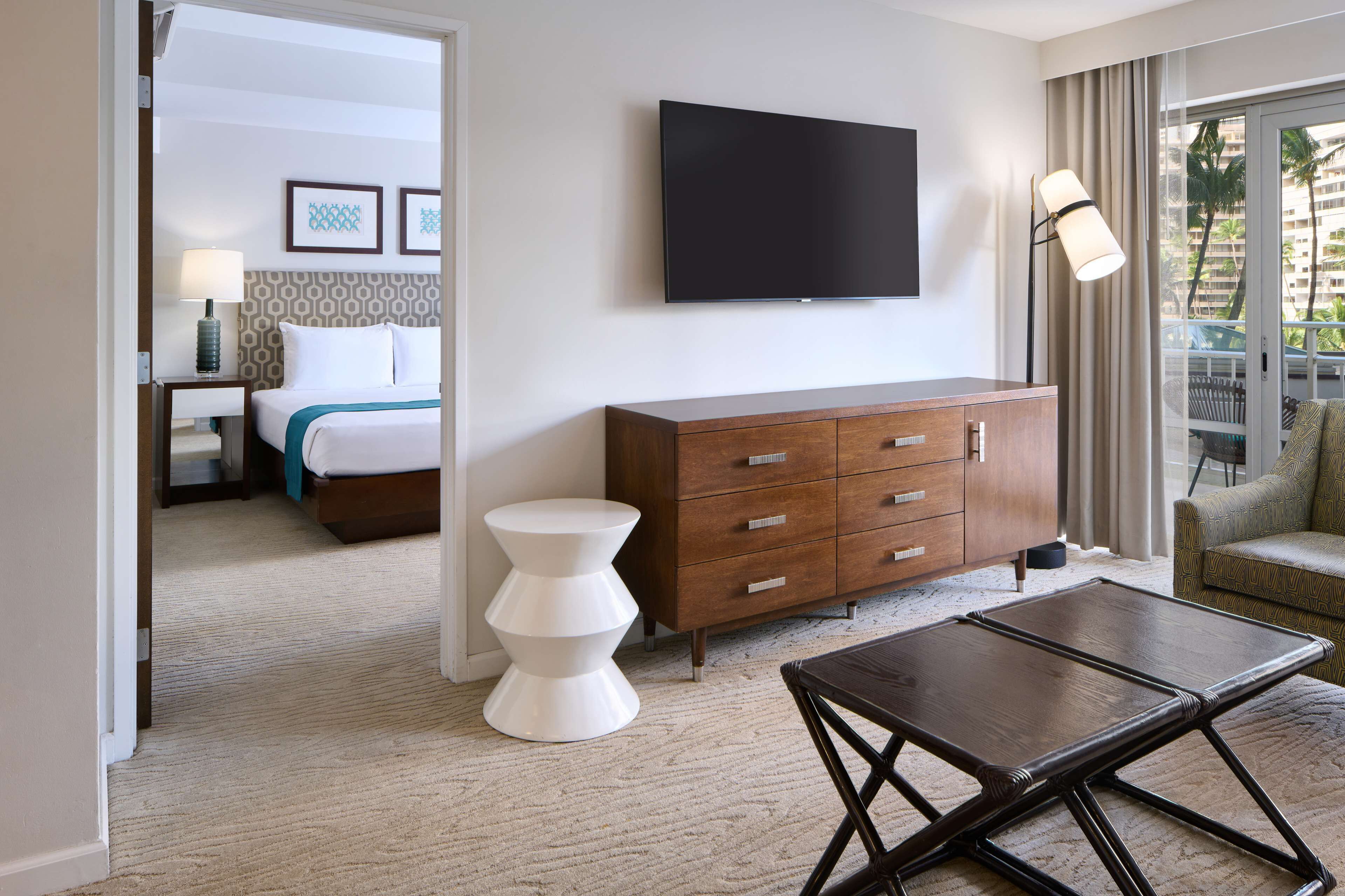 Quarto de hotel com cama, televisão, penteadeira de madeira, mesa de cabeceira branca e vista para palmeiras pela janela.