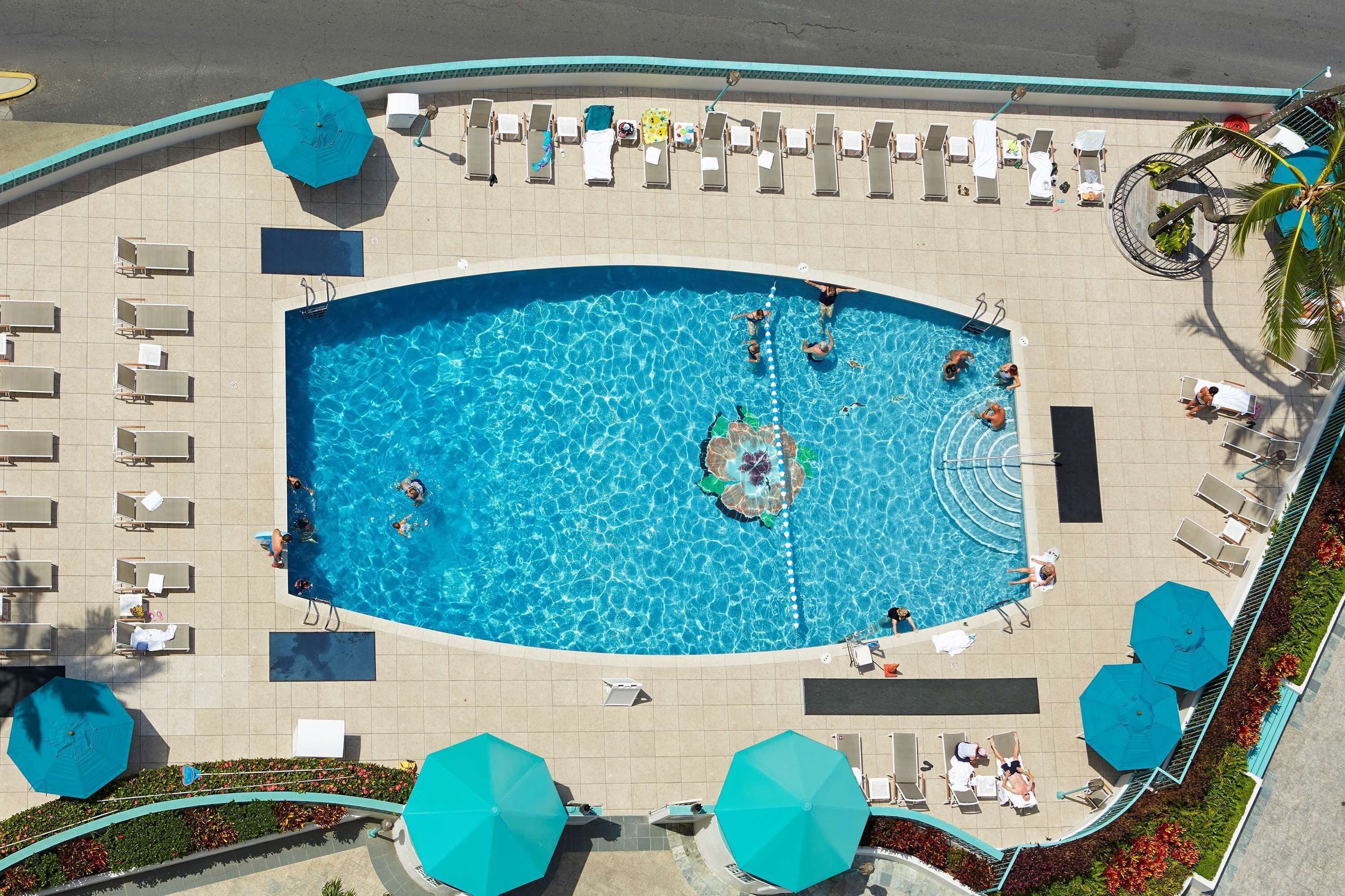 Uma piscina grande rodeada por espreguiçadeiras e guarda-sóis, com um deck ladrilhado e uma palmeira à direita.
