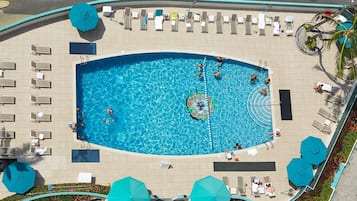 A large swimming pool surrounded by sun loungers and umbrellas, with a tiled deck and a palm tree on the right.