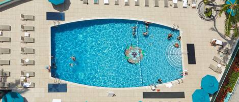 A large swimming pool surrounded by sun loungers and umbrellas, with a tiled deck and a palm tree on the right.