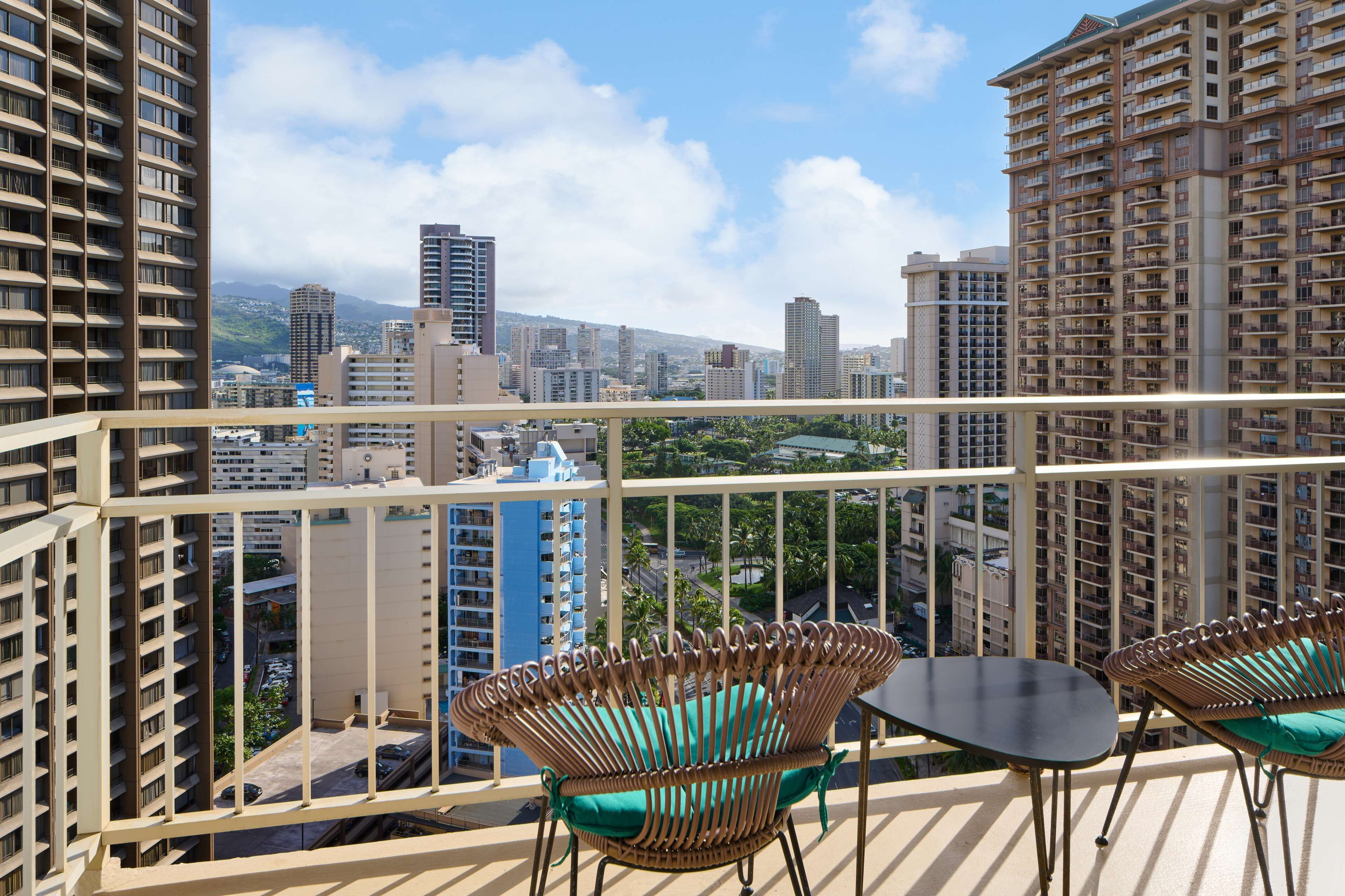 Een balkon met rieten stoelen en een tafel, met uitzicht op een stadsbeeld met hoogbouw en een heldere lucht.