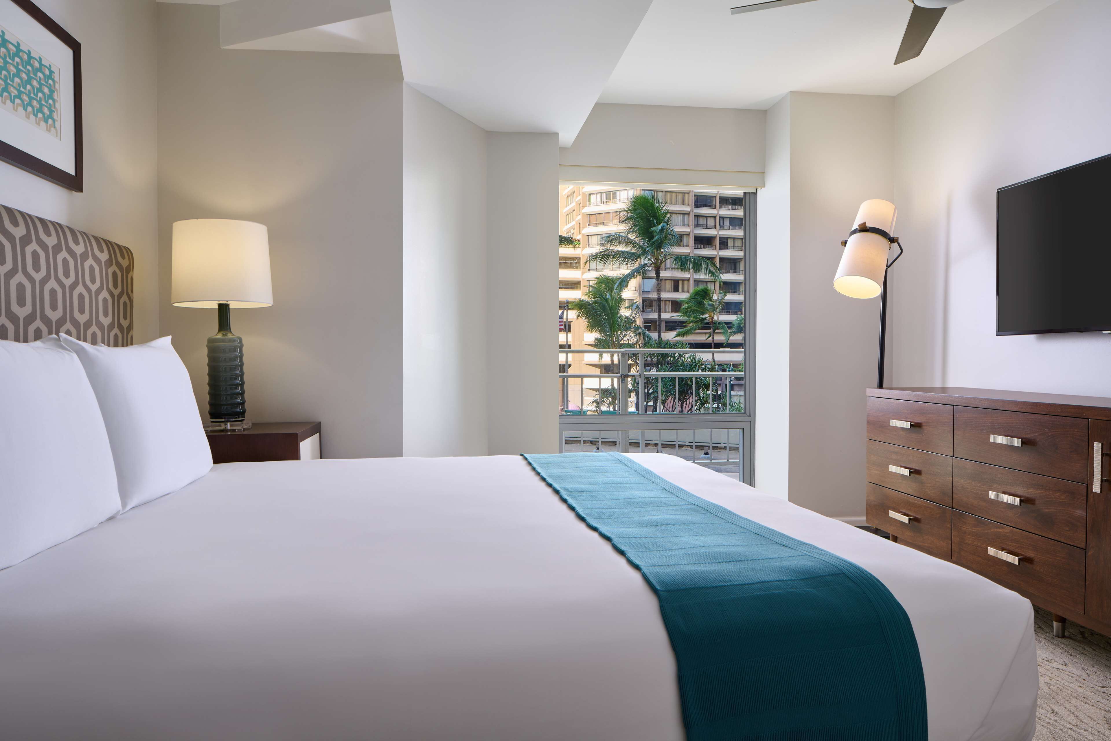 Une chambre d’hôtel comprenant un lit, une table de chevet avec une lampe, une commode et un téléviseur fixé au mur.
