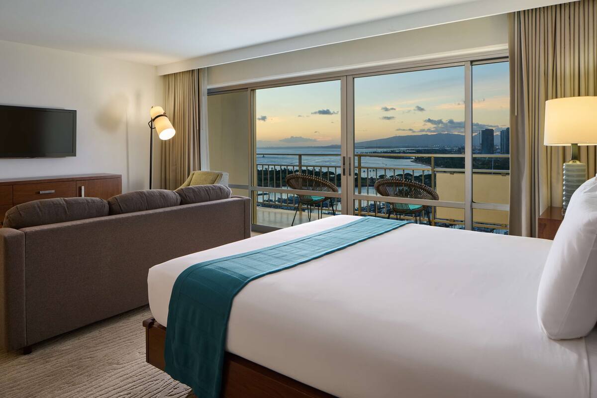 A hotel room with a large bed, a sofa, a TV, and a balcony with a view of the city and water.