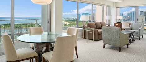 A spacious living area with a round glass table surrounded by chairs, a sofa, and a coffee table. Large windows offer a view of the city skyline and a body of water.