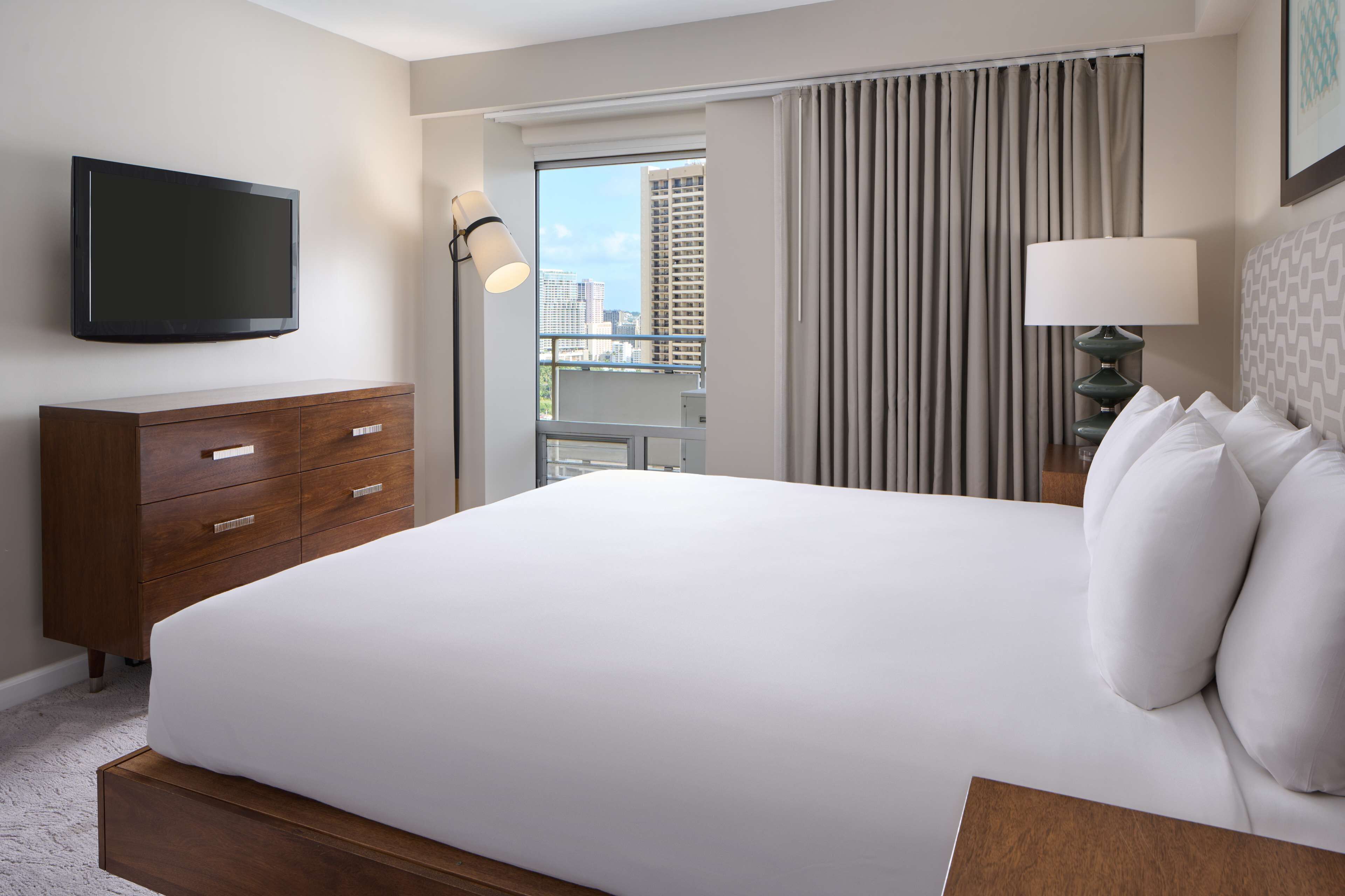 A hotel room with a large bed, a flat-screen TV mounted on the wall, a wooden dresser, a nightstand with a lamp, and a view of the city through the window.