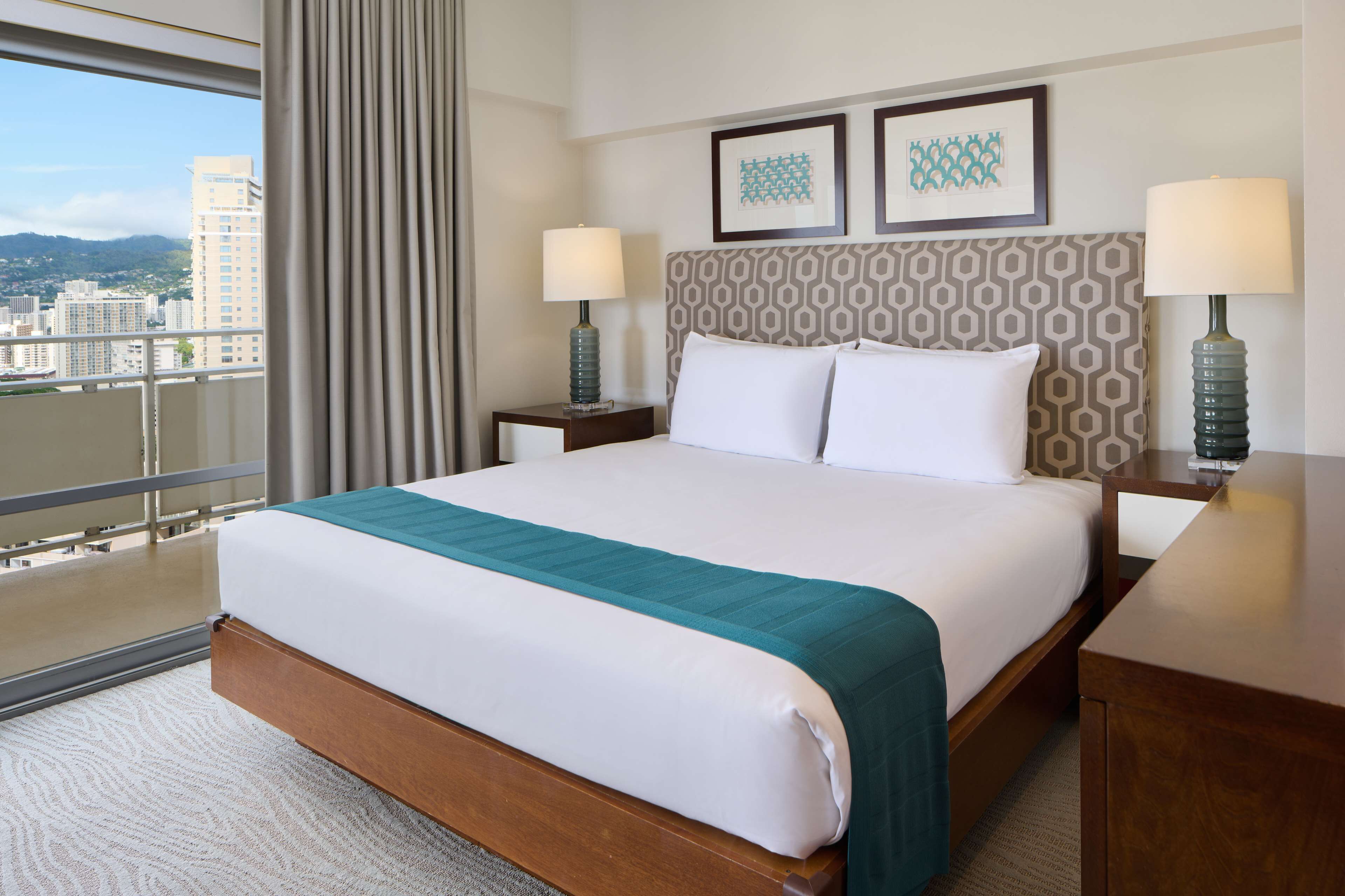 A hotel room with a large bed, two bedside tables with lamps, a view of the city through a large window, and two framed pictures on the wall.
