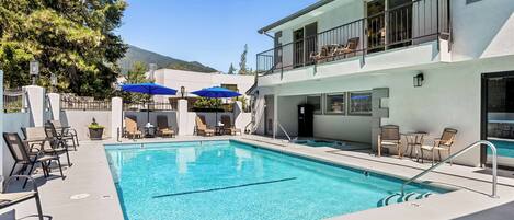 A swimming pool with lounge chairs and umbrellas, a two-story building with balconies, and a mountain in the background.