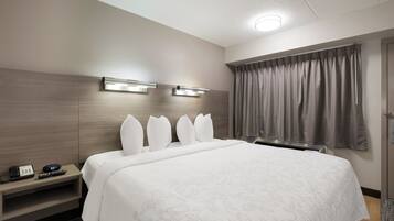 A hotel room with a large bed, a bedside table with a phone and a digital clock, a headboard with wall-mounted lights, and a window with curtains.