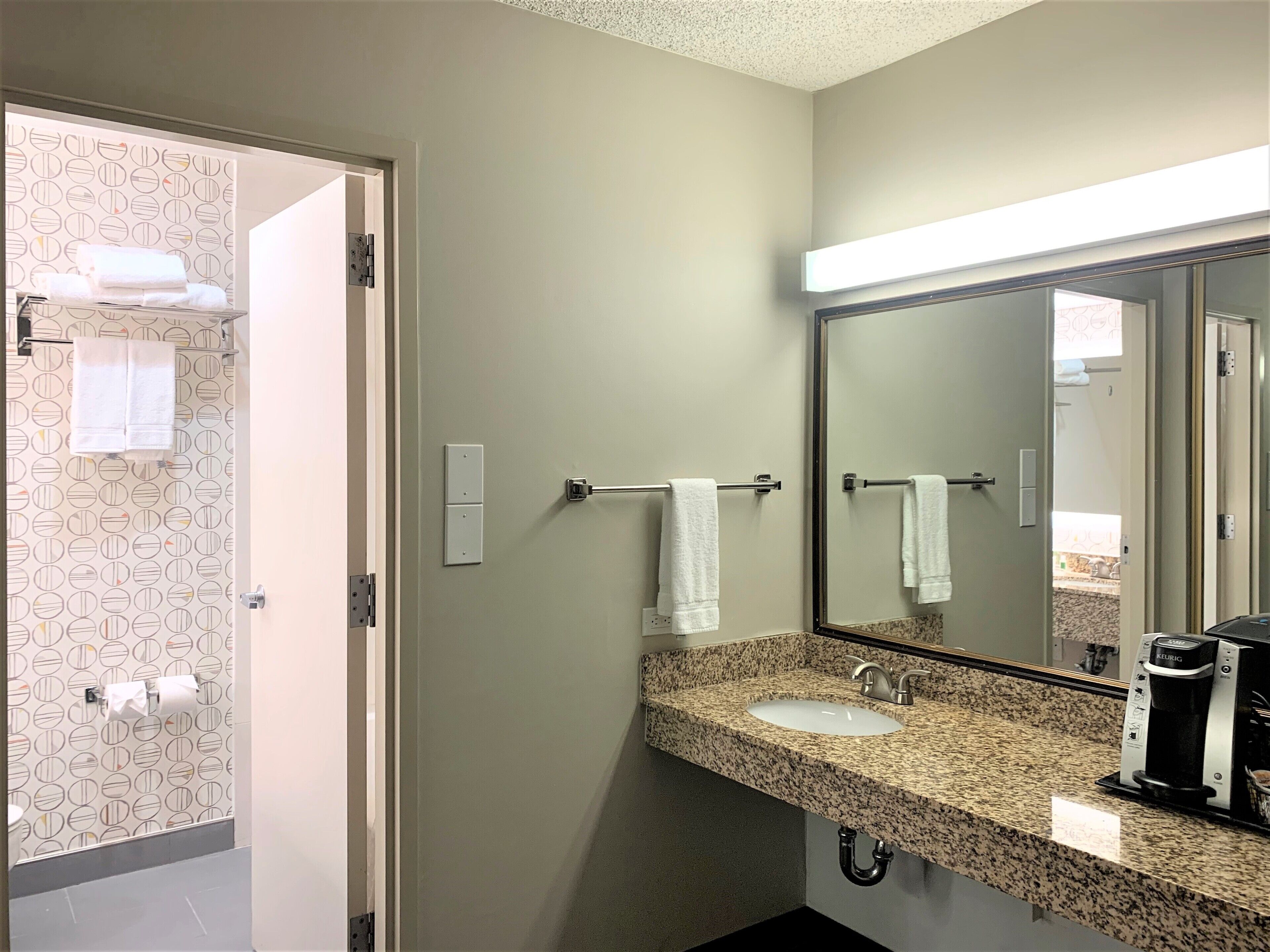 Une salle de bain avec un lavabo en granit, un miroir et une machine à café. Des serviettes sont suspendues sur un porte-serviettes et un porte-rouleau de papier toilette est fixé au mur.