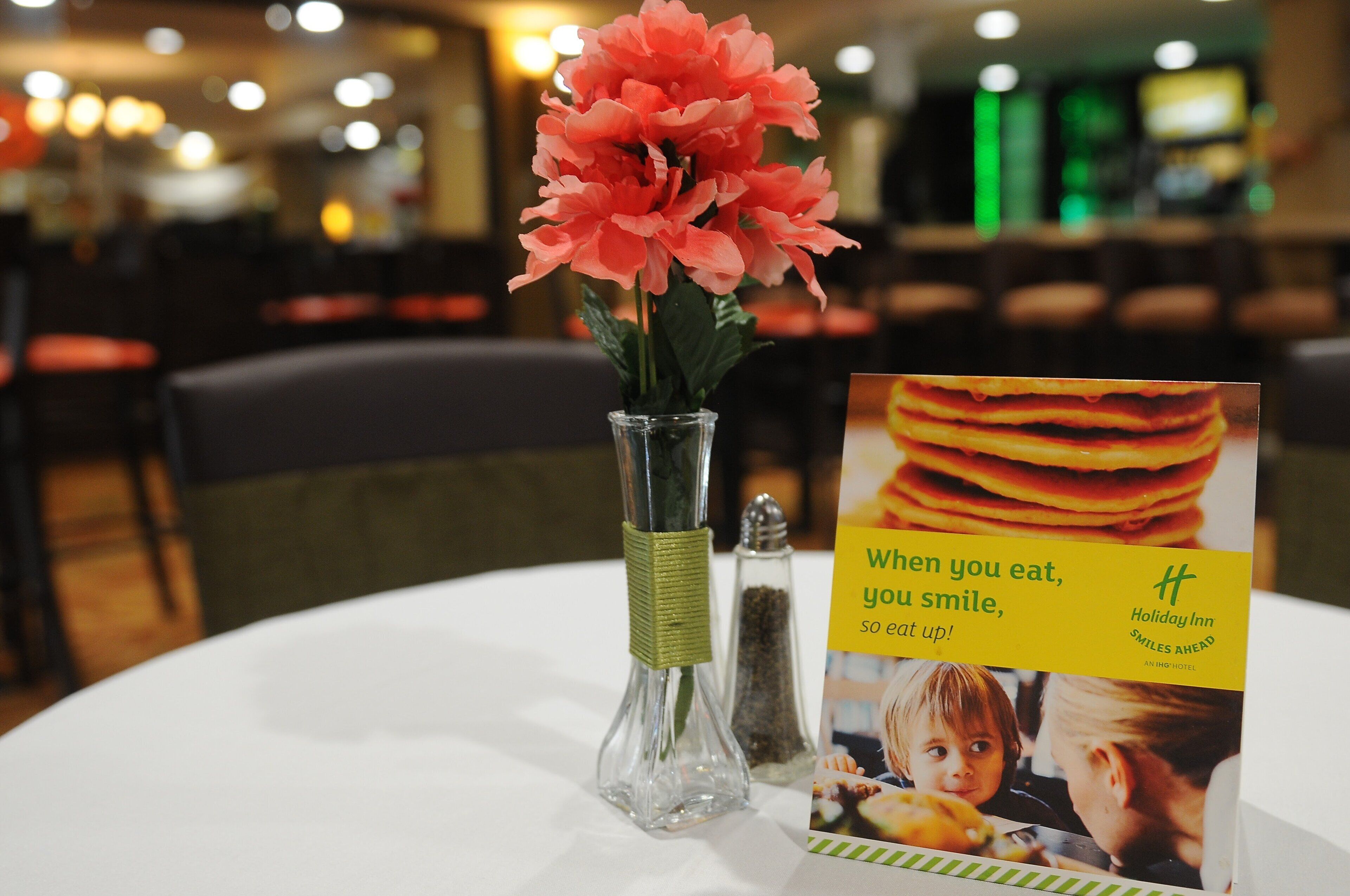 A table with a clear glass vase containing pink flowers, a salt and pepper shaker, and a promotional card with a food image and text.
