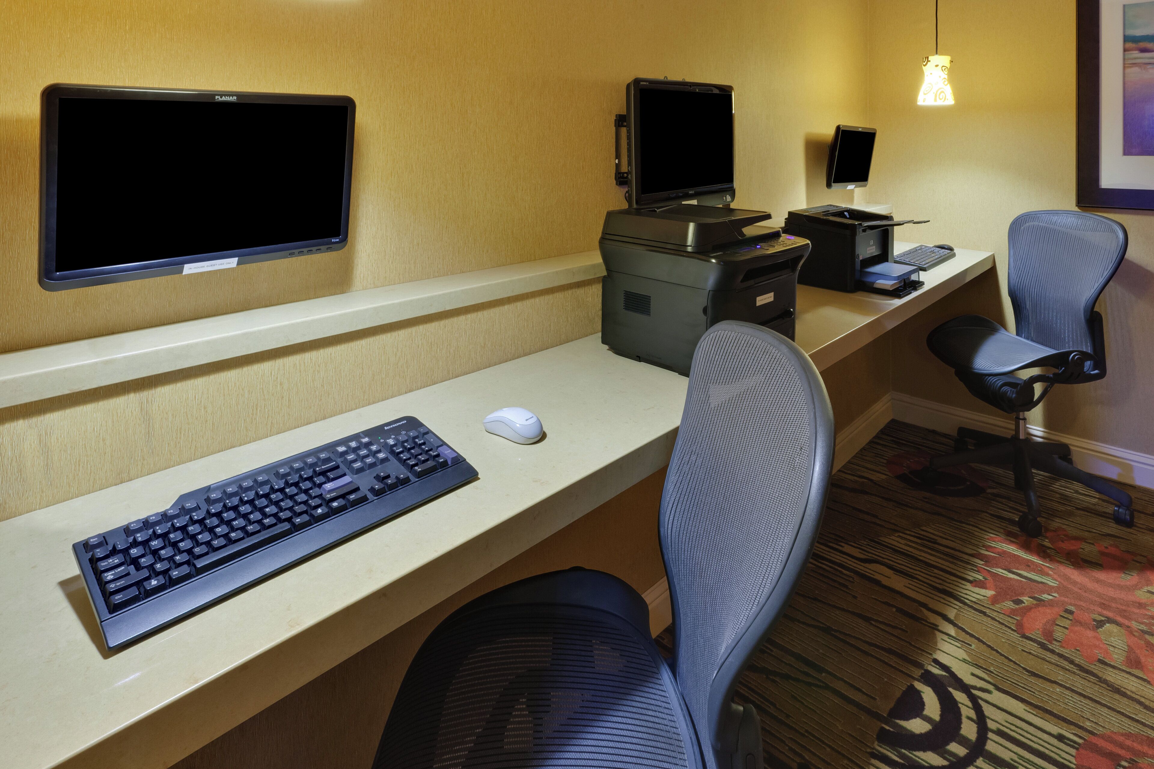A hotel room with a desk, two monitors, a printer, a keyboard, a mouse, and a chair.