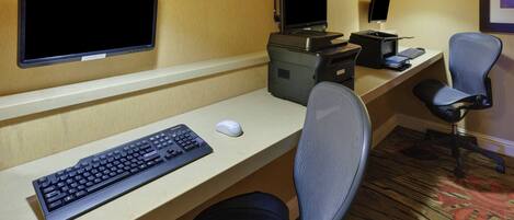 A hotel room with a desk, two monitors, a printer, a keyboard, a mouse, and a chair.