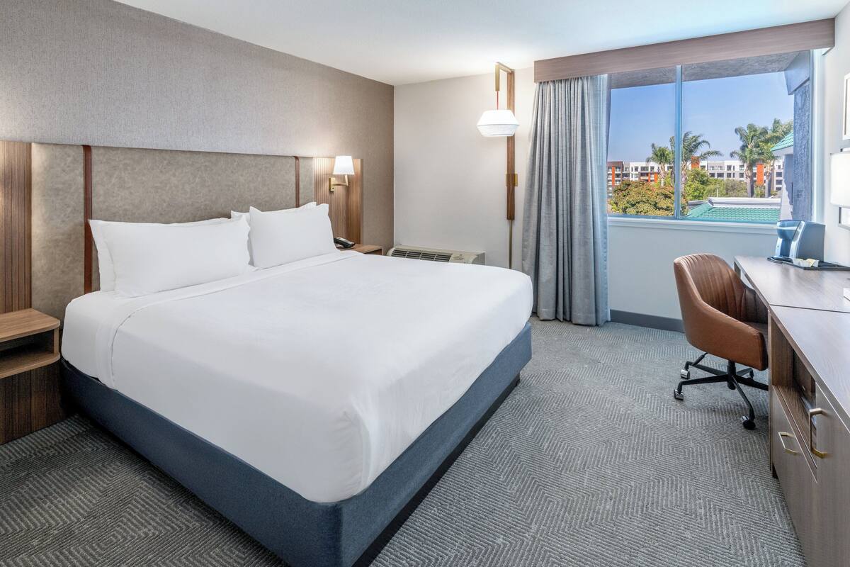 A hotel room with a large bed, a desk, a chair, a desk lamp, a nightstand, and a window with a view of buildings and palm trees.
