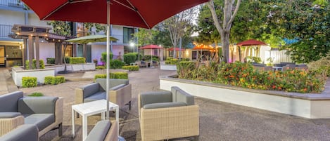 Un patio al aire libre con mobiliario de mimbre, sombrillas rojas y un jardín cuidado.