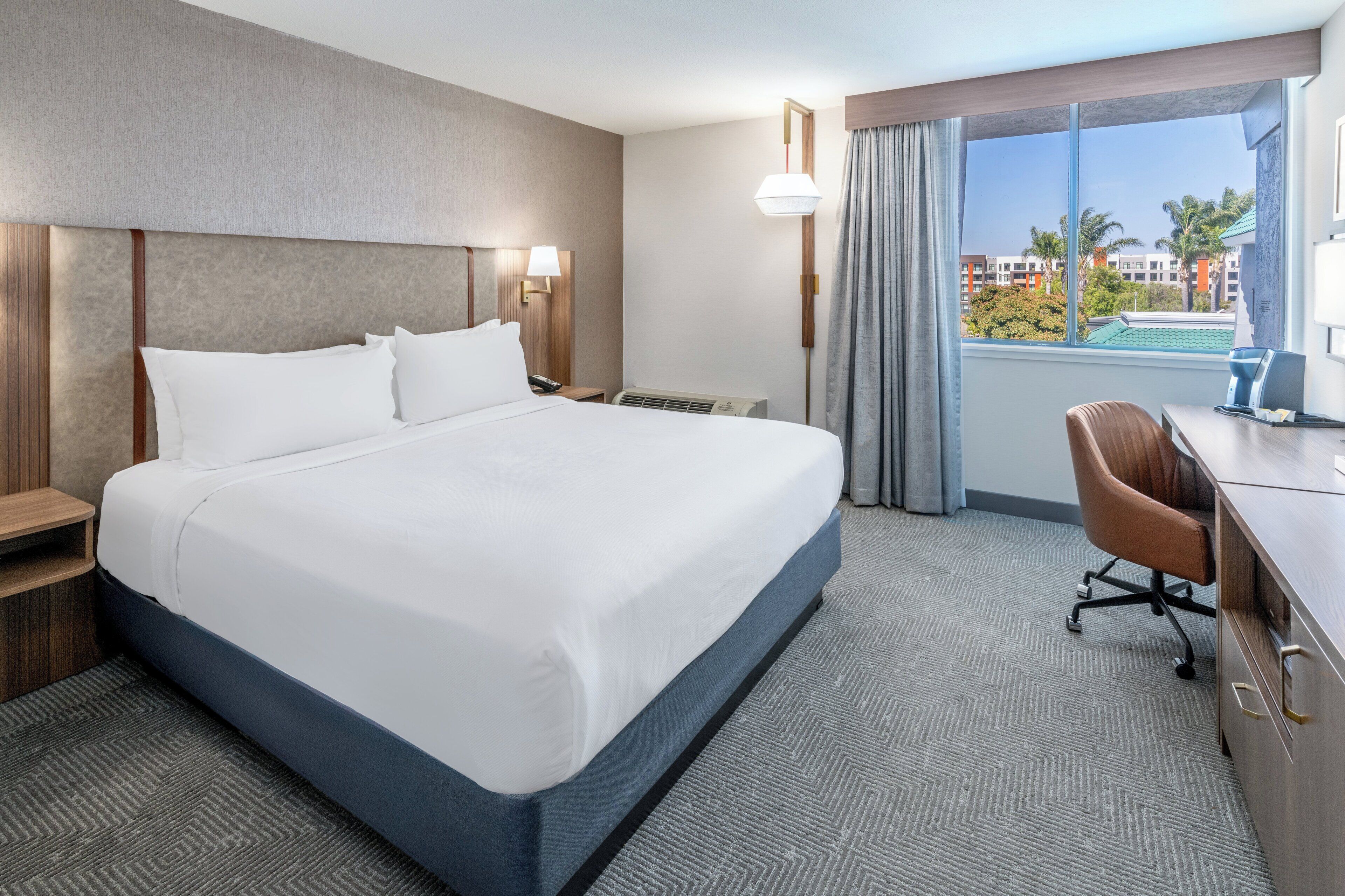 A hotel room with a large bed, a desk, a chair, a desk lamp, a nightstand, and a window with a view of buildings and palm trees.