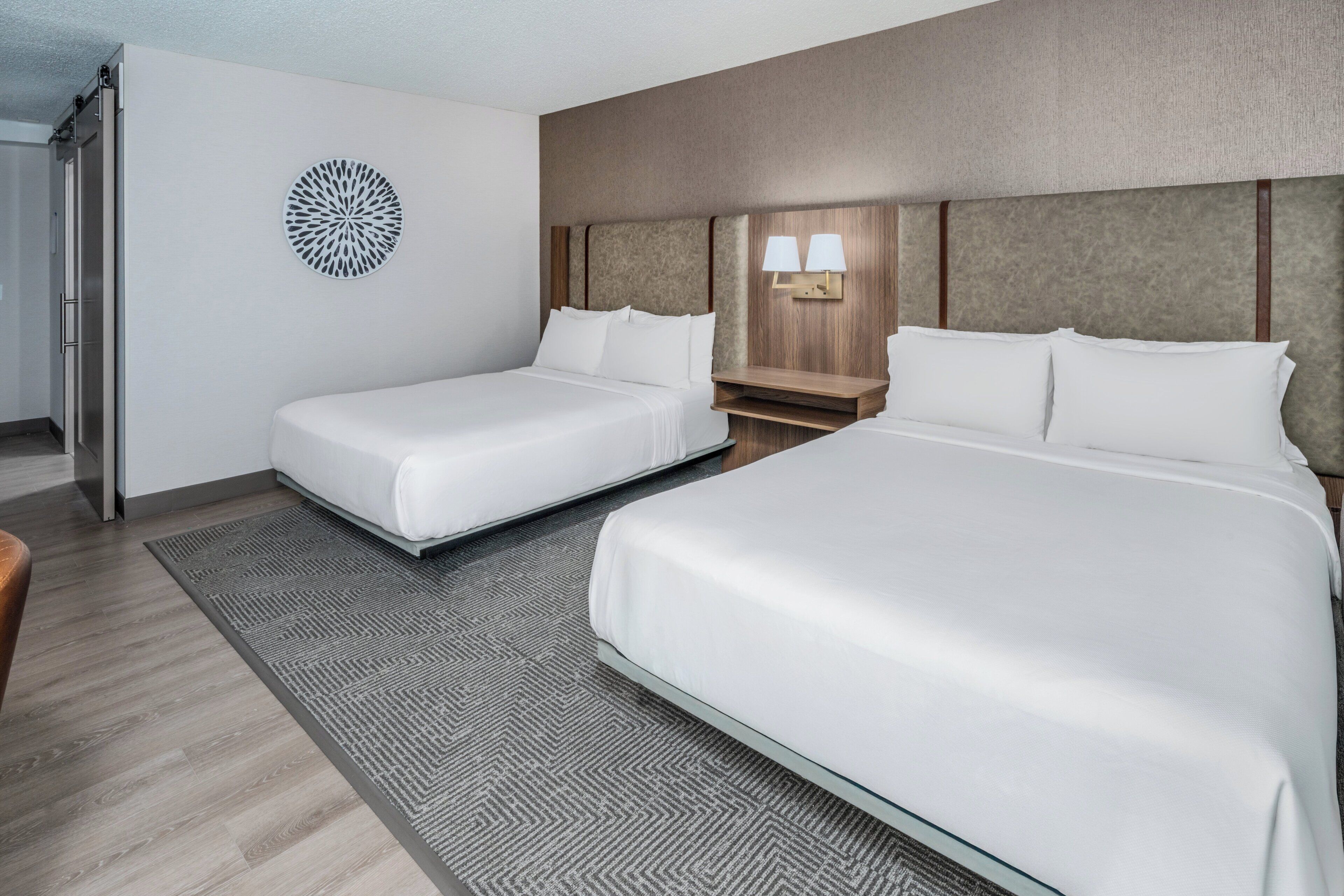 A hotel room with two beds, a small table, a wall-mounted lamp, and a decorative circular piece on the wall.