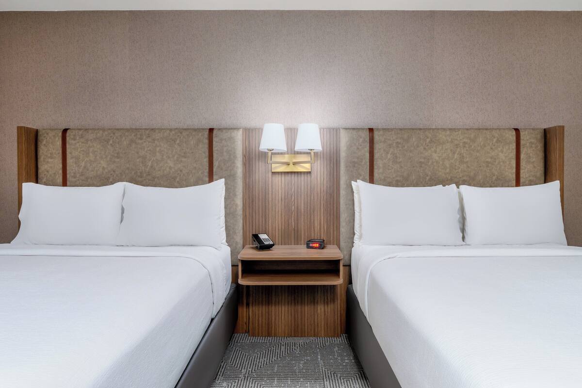 A hotel room with two beds, a nightstand with a phone and alarm clock, and a wall-mounted lamp.