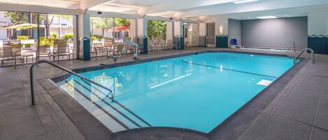 An indoor swimming pool with clear water, surrounded by lounge chairs and tables, and large windows offering views of the outside.