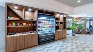 A modern hotel lobby with a vending machine stocked with snacks and drinks, wooden shelving with various items, and a potted plant. - Holiday Inn Dublin-Pleasanton by IHG (Dublin)