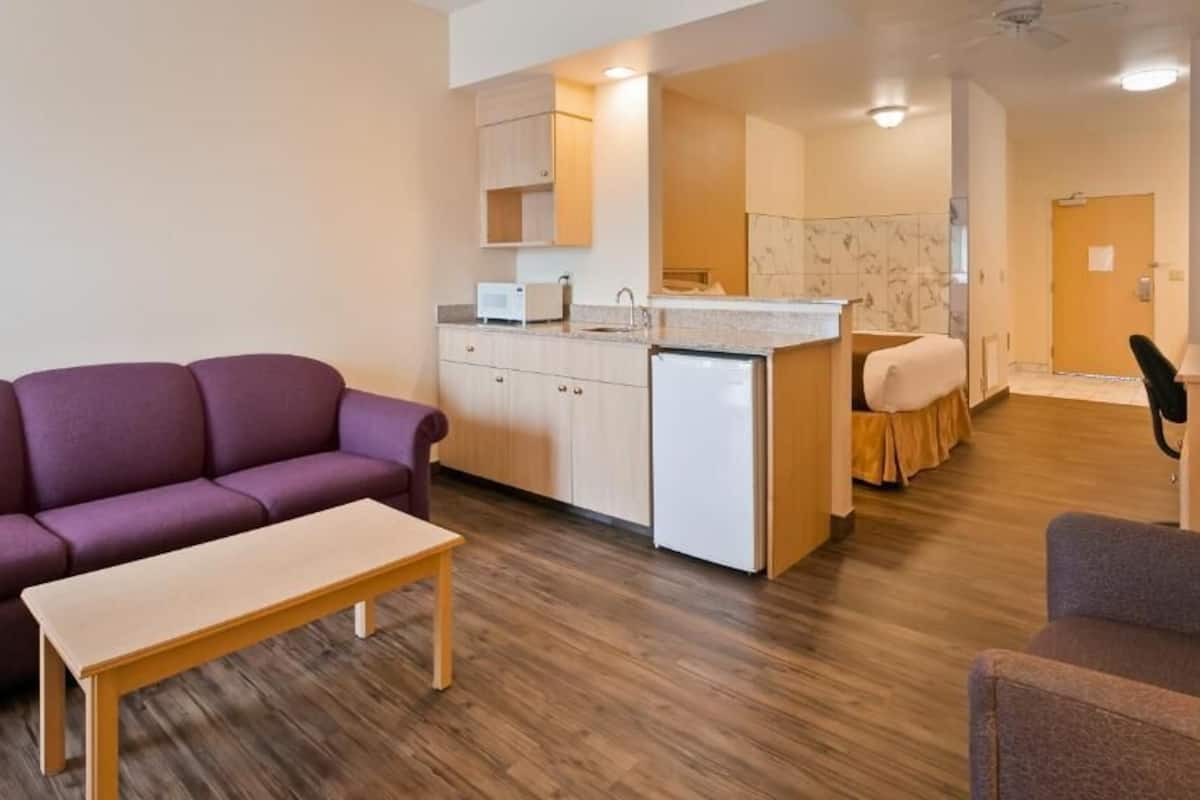 A compact living space with a purple sofa, a wooden table, a kitchenette, a small bed, and a television.