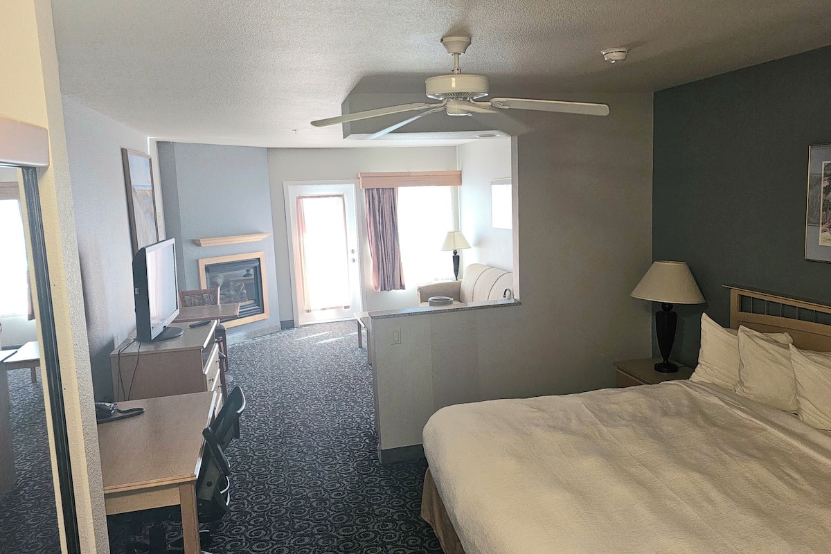 A hotel room with a bed, a desk with a chair, a ceiling fan, a fireplace, a window with curtains, and a framed picture on the wall.