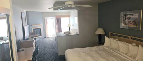 A hotel room with a bed, a desk with a chair, a ceiling fan, a fireplace, a window with curtains, and a framed picture on the wall.