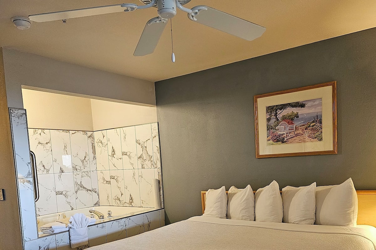 A large bed with a headboard, a ceiling fan, a painting on the wall, and a bathroom with a marble-tiled vanity.
