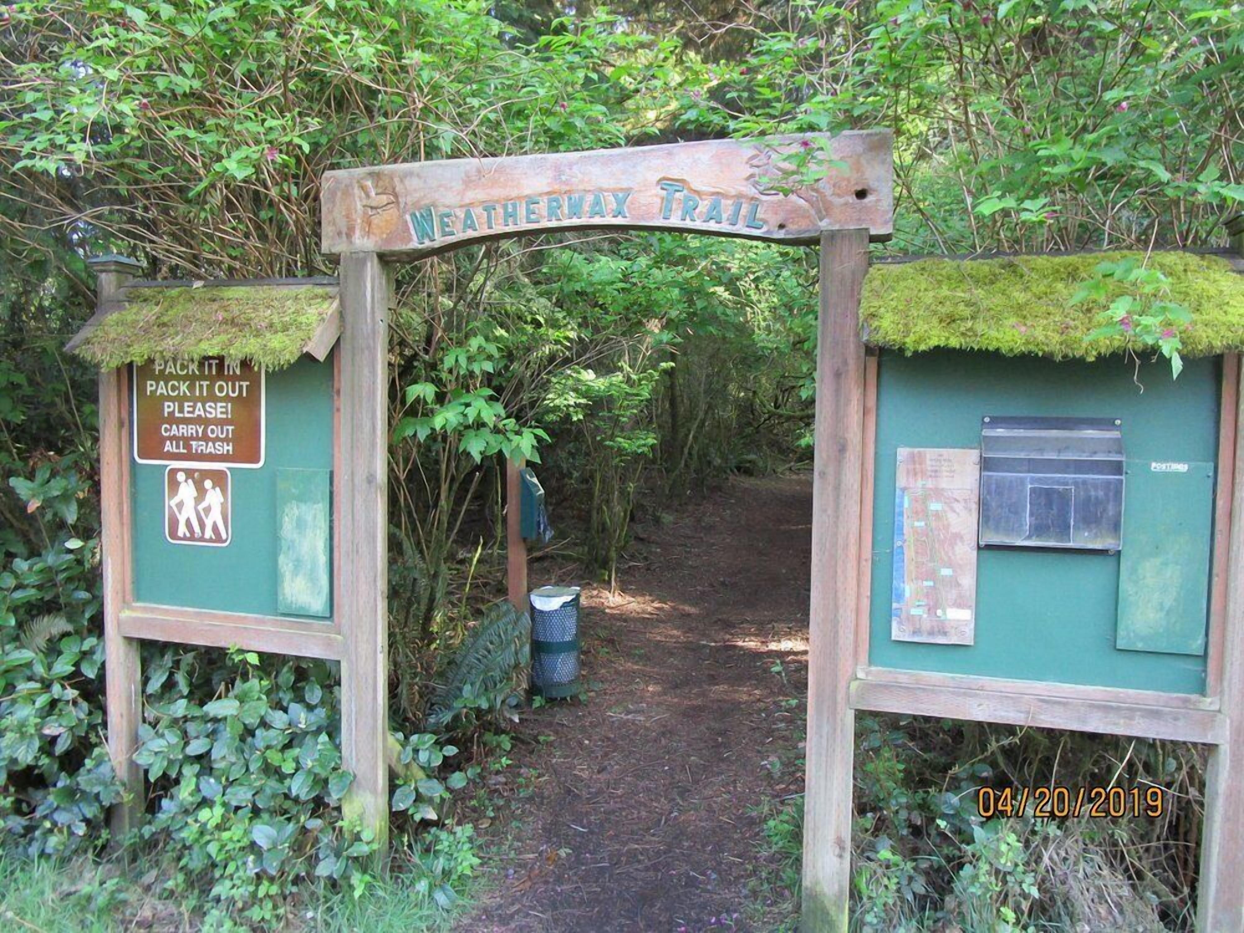 A wooden sign for Weatherwax Trail with a trash can and a recycling bin.