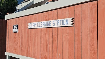 Estructura de madera con el rótulo "CLAM CLEANING STATION" y un letrero que dice "NO FUMAR", junto a un edificio con la señalización "Lighthouse Suites Inn".