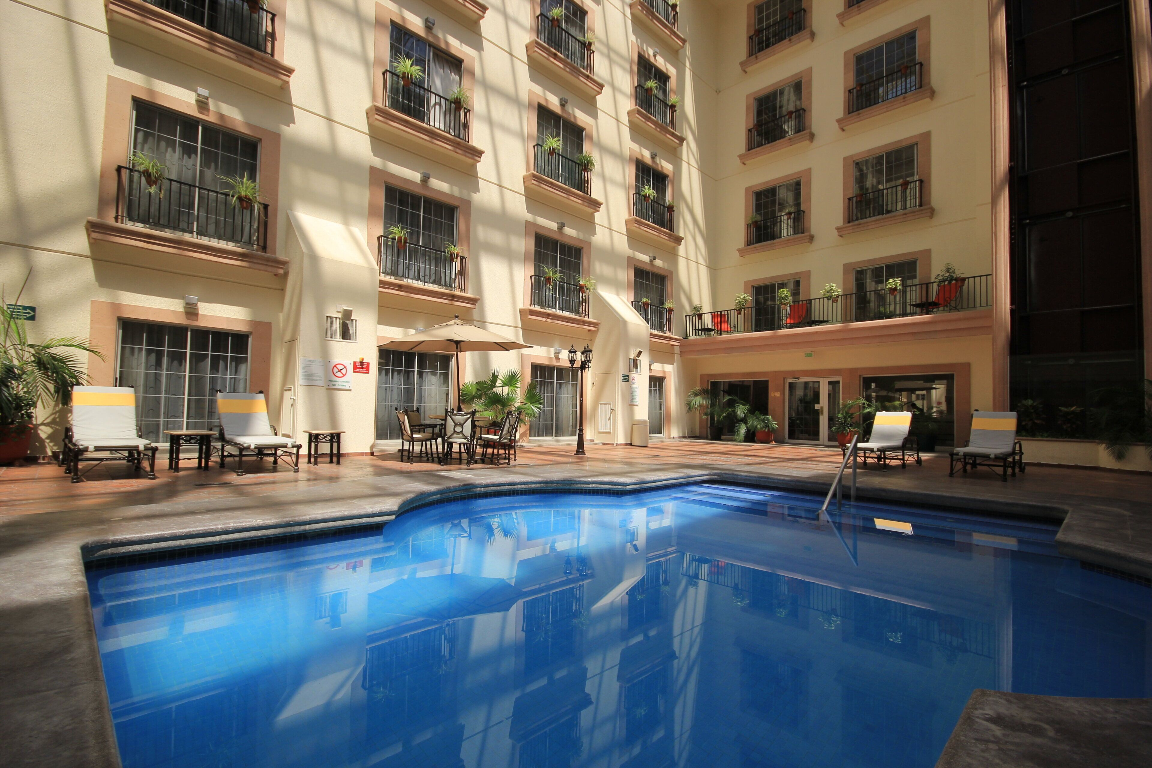 A hotel pool area with multiple lounge chairs and tables, surrounded by balconies with potted plants.