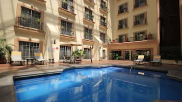 A hotel pool area with multiple lounge chairs and tables, surrounded by balconies with potted plants.