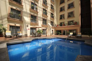 A hotel pool area with multiple lounge chairs and tables, surrounded by balconies with potted plants.