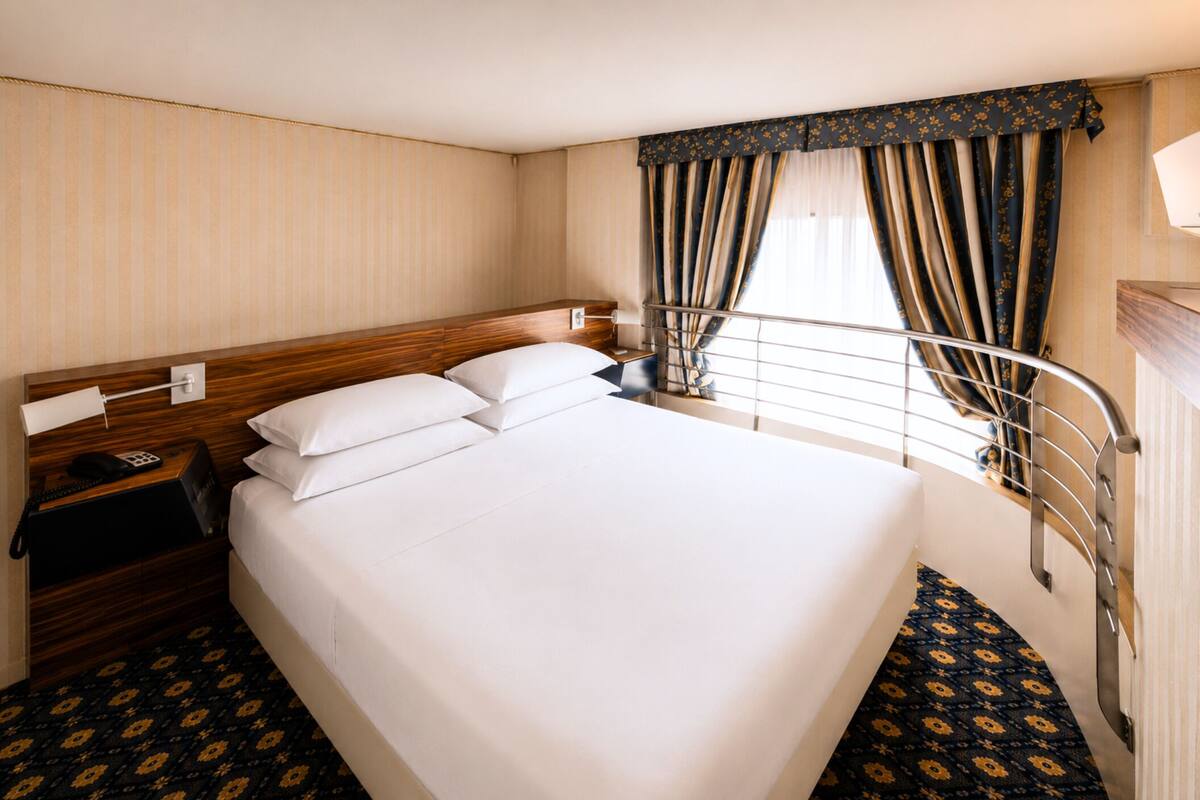 A hotel room with a large bed, a wooden headboard, a nightstand with a phone, and a window with curtains.