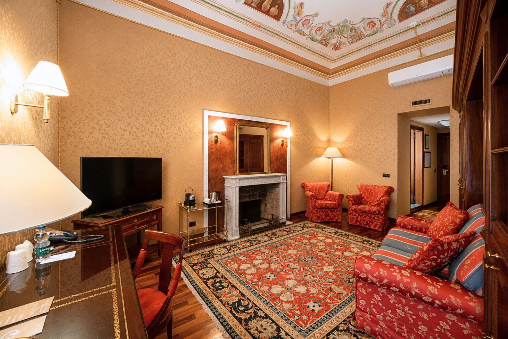 Una habitación de hotel con televisión, chimenea, un sofá rojo, alfombra estampada y una mesa de comedor con sillas.