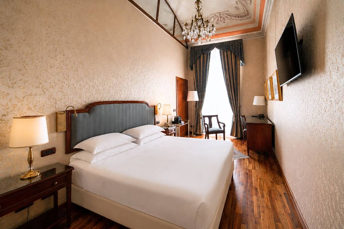 A hotel room with a large bed, a desk with a lamp, a TV mounted on the wall, and a chandelier.