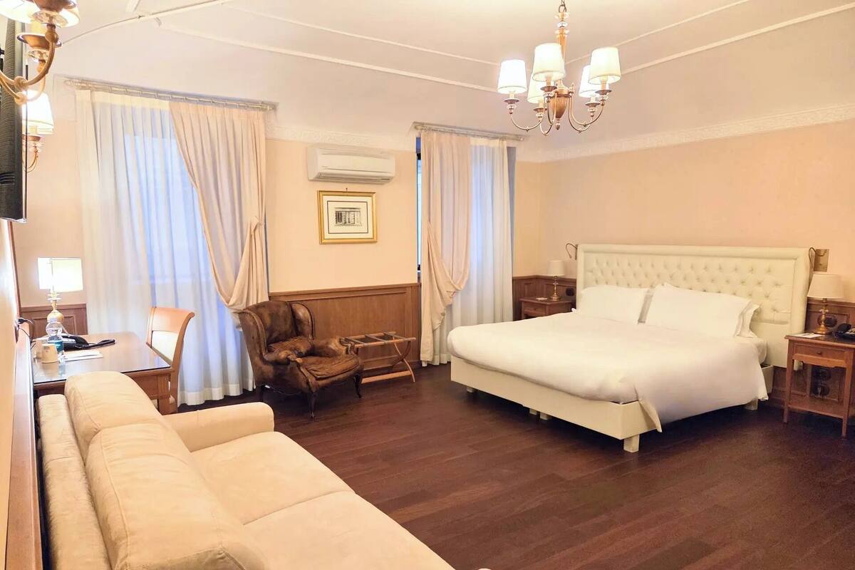 A hotel room with a large bed, a sofa, a chair, a desk with a lamp, and a chandelier.